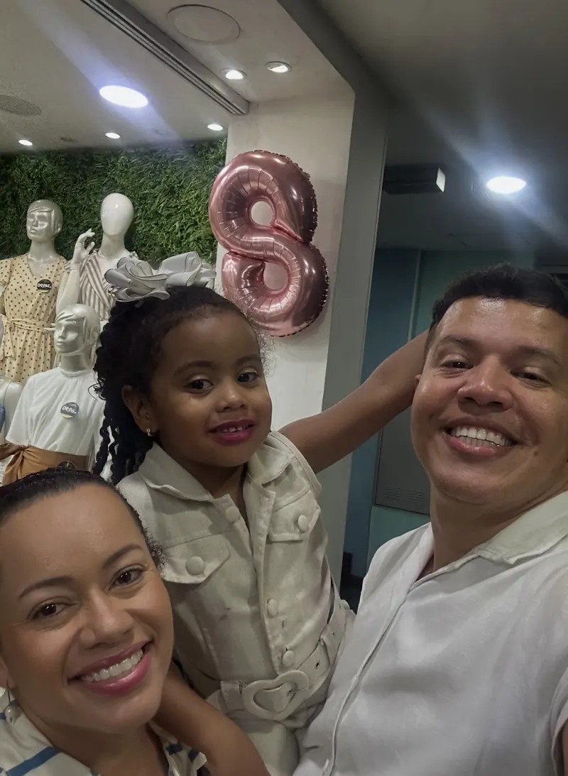 Os donos da San Maria sorrindo com a filha no colo, dentro da loja, com o balão de 8 anos e os manequins ao fundo.
